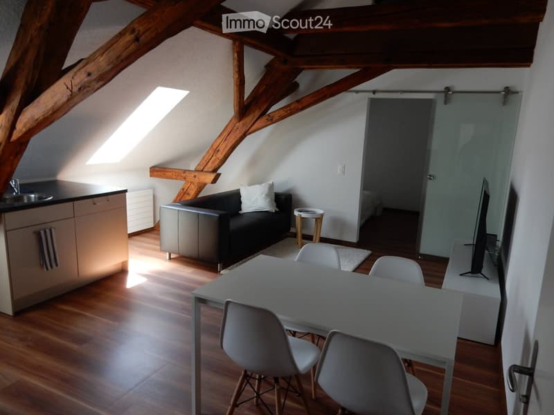 Open-plan living and dining area with exposed wooden beams, a sofa, a dining table with chairs, a small kitchen area, and a TV screen.
