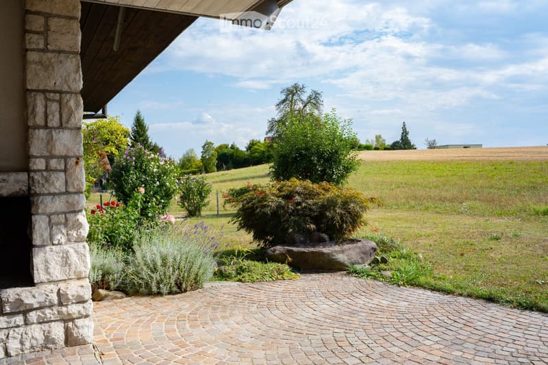 Garden area with a paved pathway, stone pillar, and various plants and flowers, leading to a grassy field with trees and a clear sky.