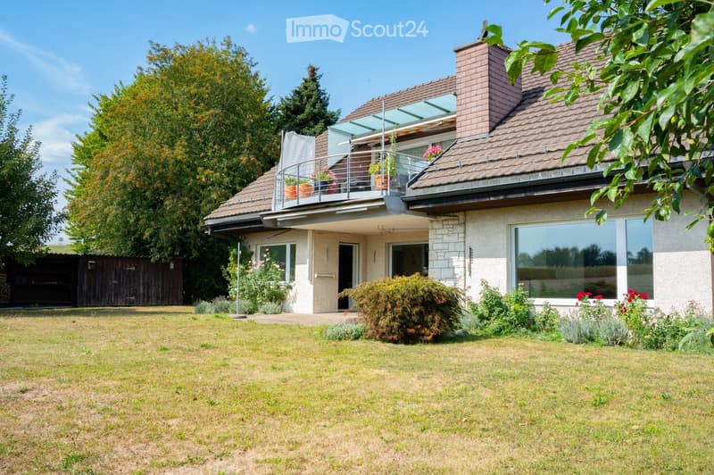Two-story house with a tiled roof, large windows, and a balcony on the upper level. The house is surrounded by a well-maintained lawn and garden with various plants and flowers.
