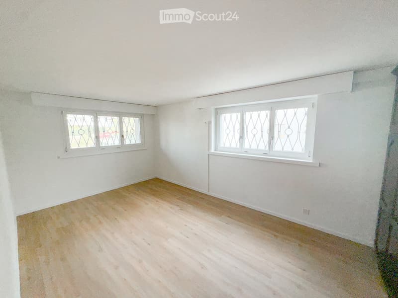 Empty room with light wooden flooring and white walls, featuring two windows with decorative grilles.