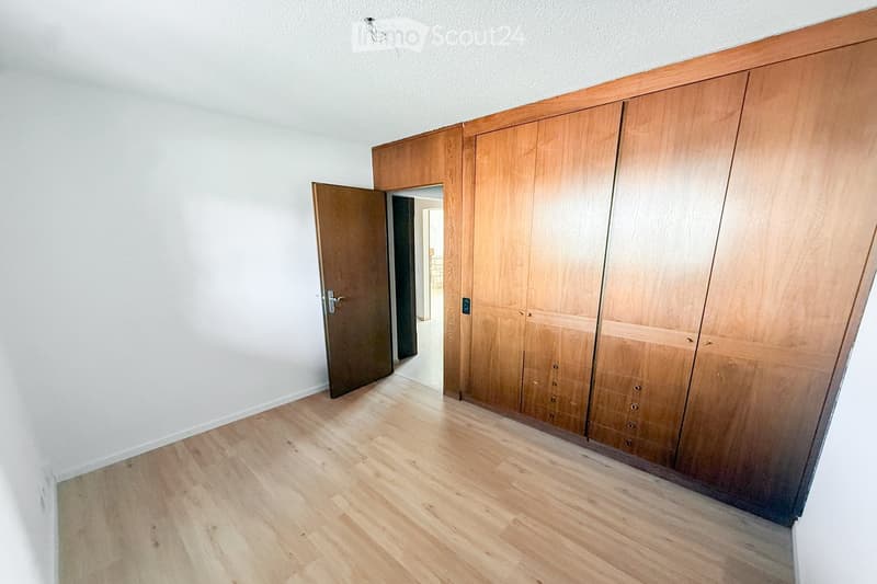 Empty room with light wooden flooring, white walls, and built-in wooden wardrobes. A door is open, revealing a glimpse of another room.