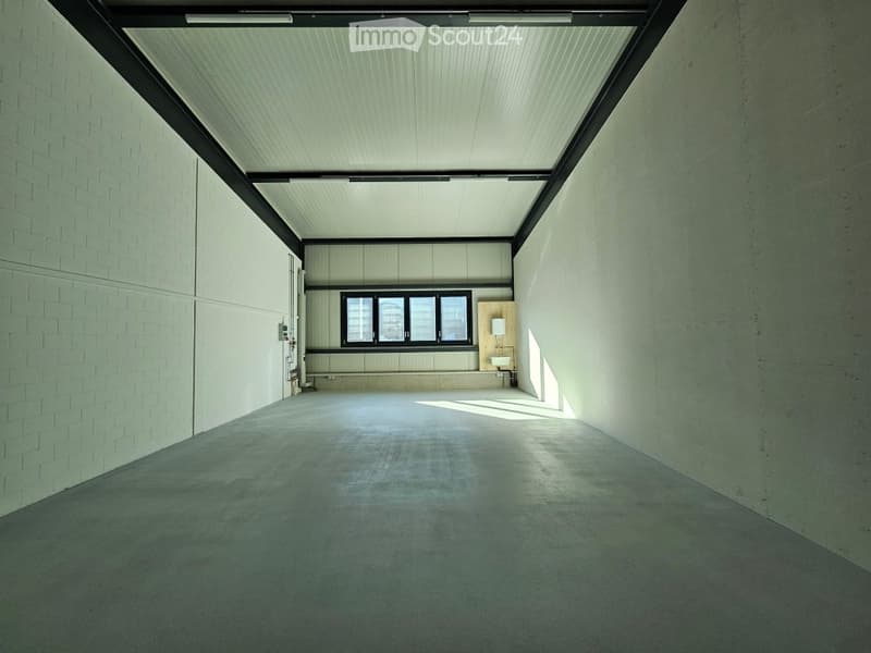 Empty room with concrete floor, white brick walls, and a large window at the far end. The ceiling is white with a black frame and linear lighting fixtures.