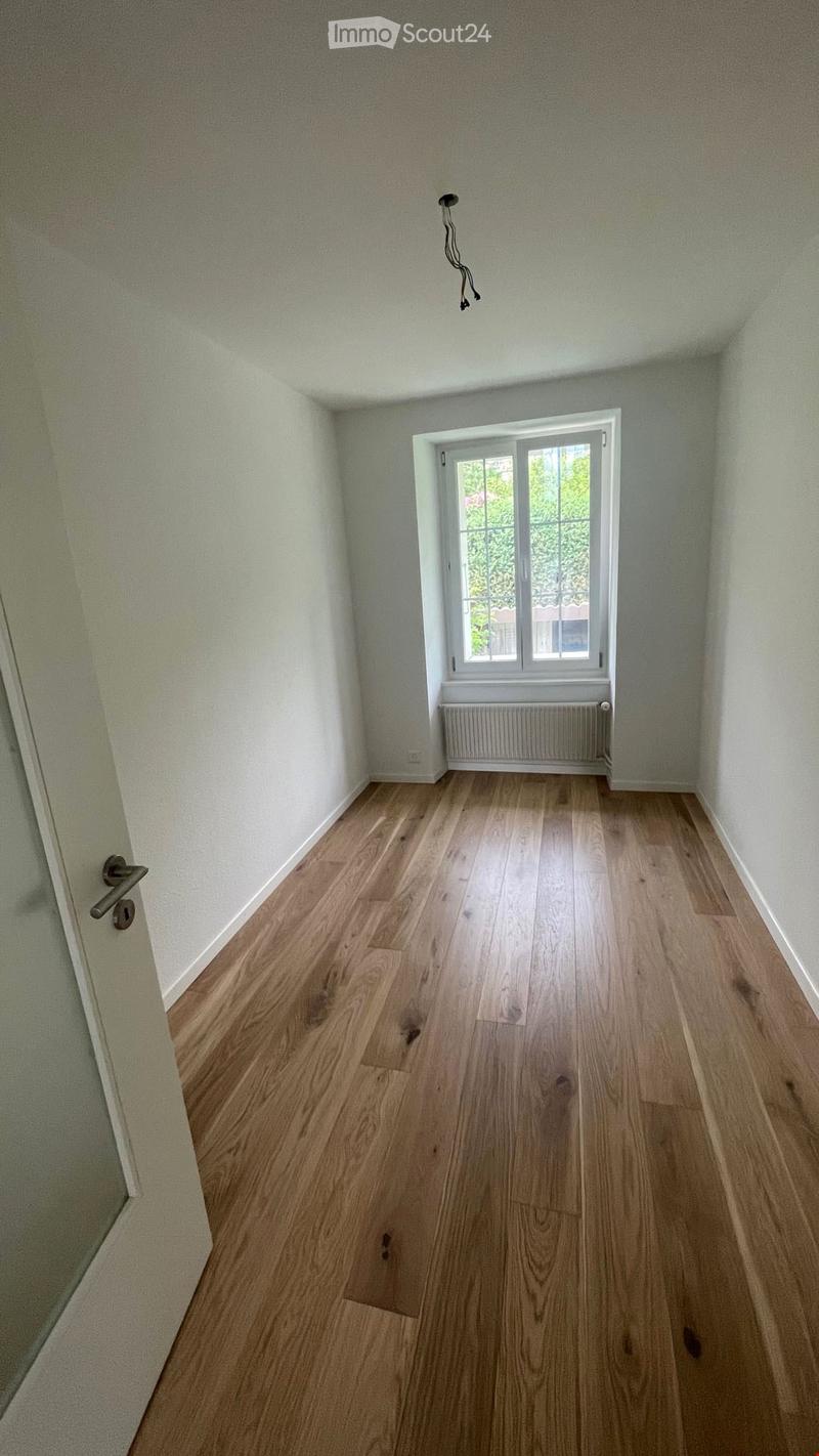 Empty room with light wooden flooring, white walls, a window with white frame, and a radiator below the window. A door is visible on the left side.