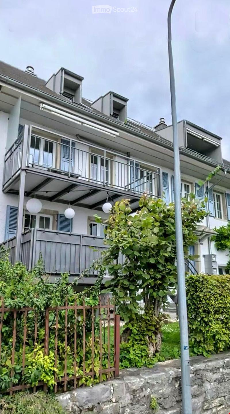 Multi-story residential building with balconies, grey exterior, and lush greenery in front.