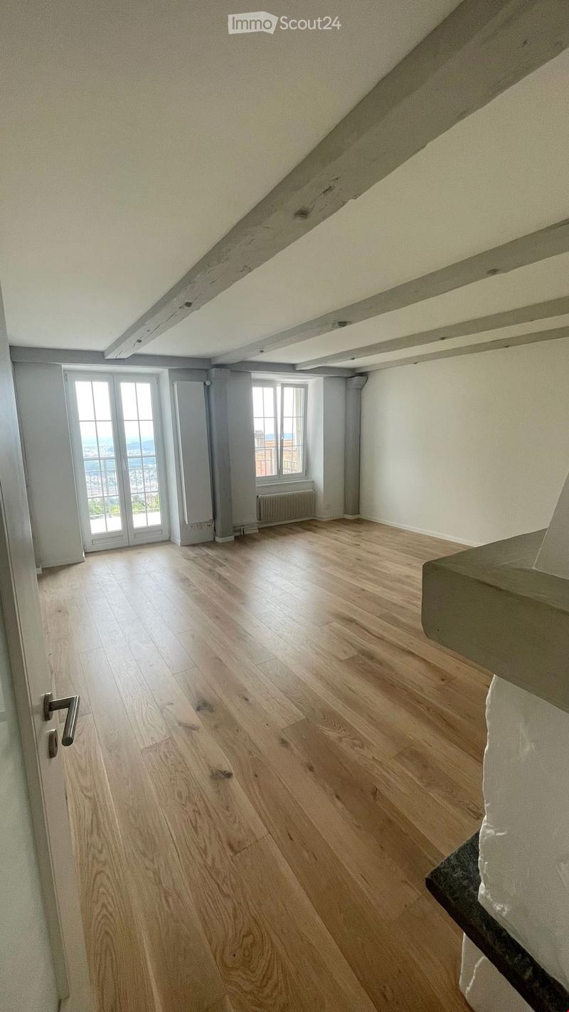 Empty room with wooden flooring, white walls, large windows, and a radiator. The ceiling has exposed wooden beams.