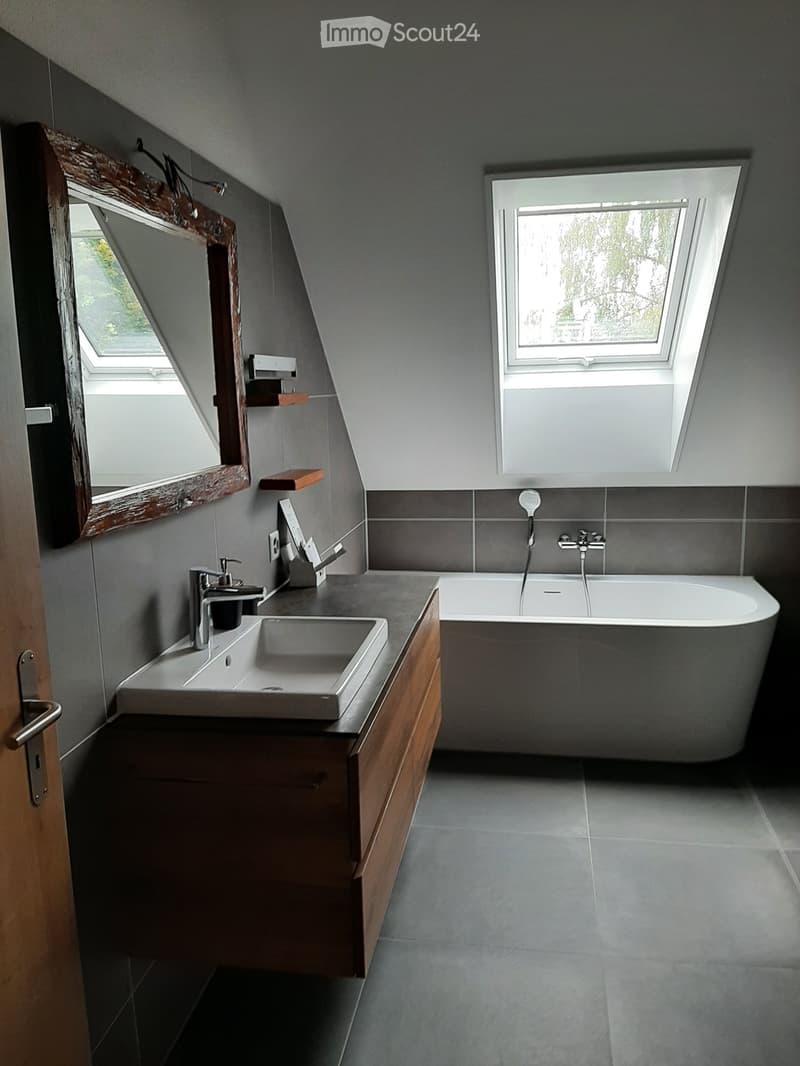 Modern bathroom with a double sink, wooden cabinet, bathtub, and large mirror. Features skylight windows and sleek grey tiles.