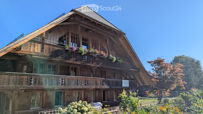 Traditional wooden house with multiple stories, balconies, and flower boxes, surrounded by greenery and a clear blue sky.