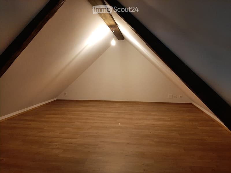 Empty attic space with wooden flooring, slanted ceilings, and exposed wooden beams. A single light fixture hangs from the ceiling.