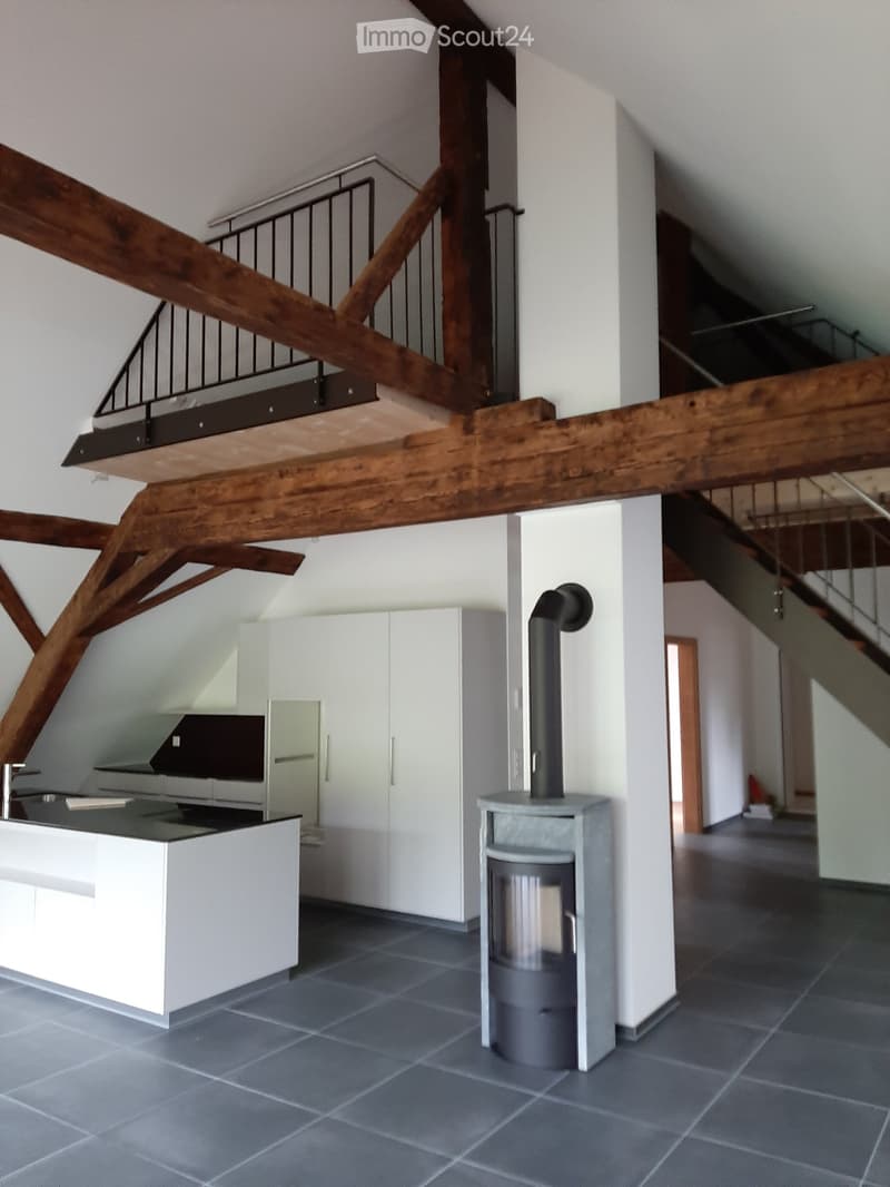 Modern kitchen area with white cabinets, a kitchen island, and a wood stove, featuring exposed wooden beams and a staircase leading to an upper level.