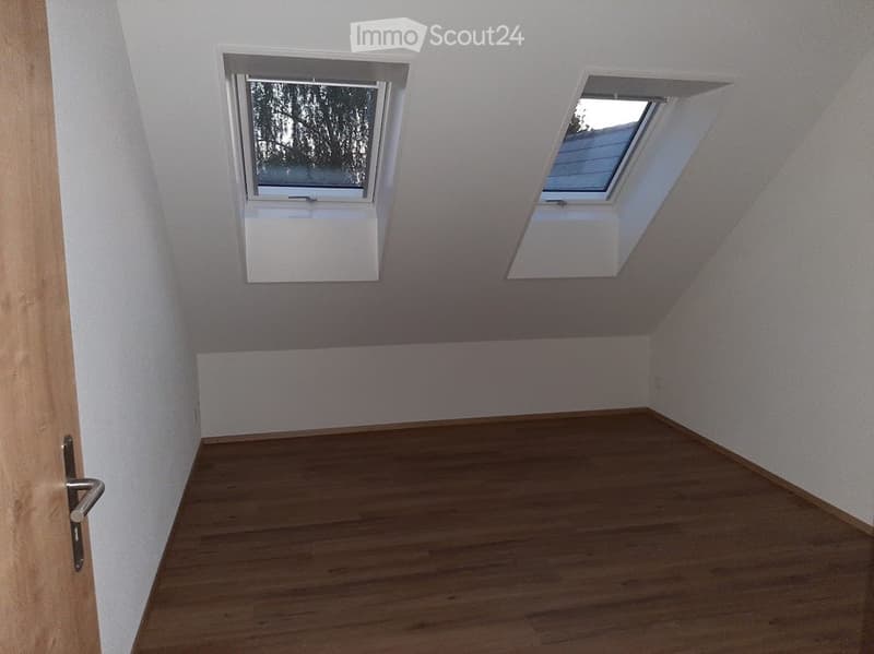 Empty attic room with wooden flooring, white walls, and two windows, one of which is a skylight.