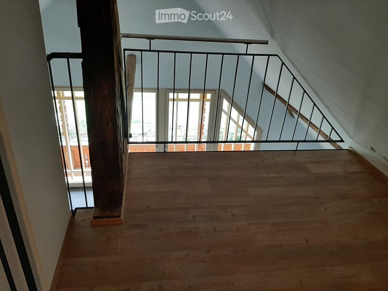 Wooden staircase with metal railing, leading to a bright room with large windows and wooden furniture visible in the background.