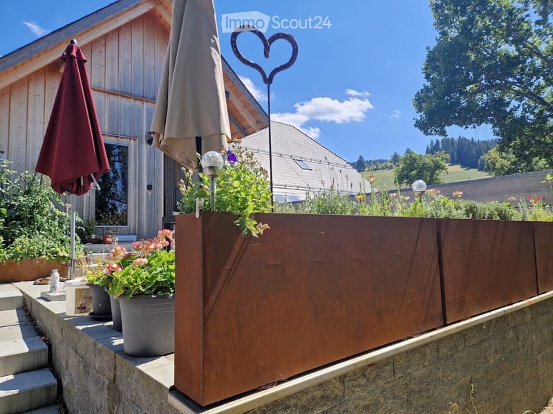 Outdoor garden area with wooden planter, potted plants, and patio umbrellas near a wooden house.