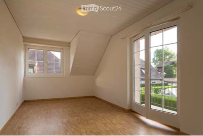 Empty attic room with wooden flooring, white walls, and large windows including a sliding glass door leading to a balcony.