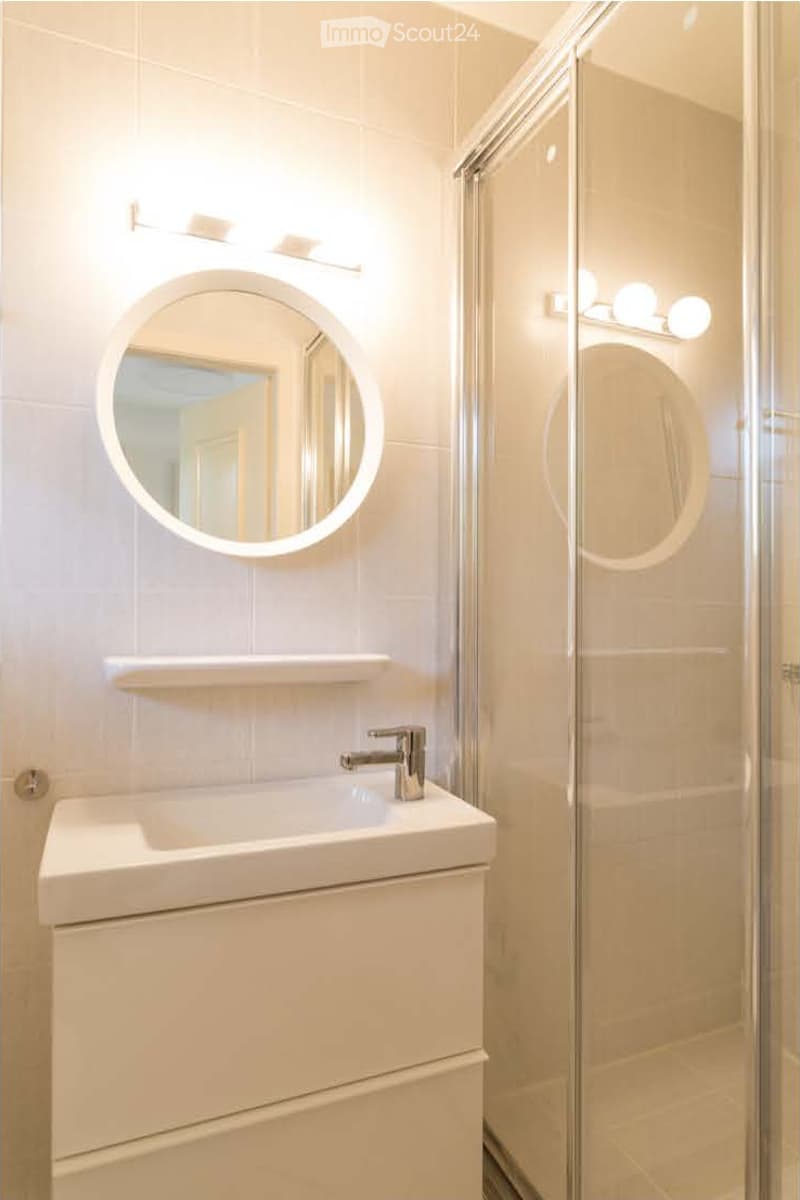 Modern bathroom with a white sink, chrome faucet, round illuminated mirror, and a glass-enclosed shower area.