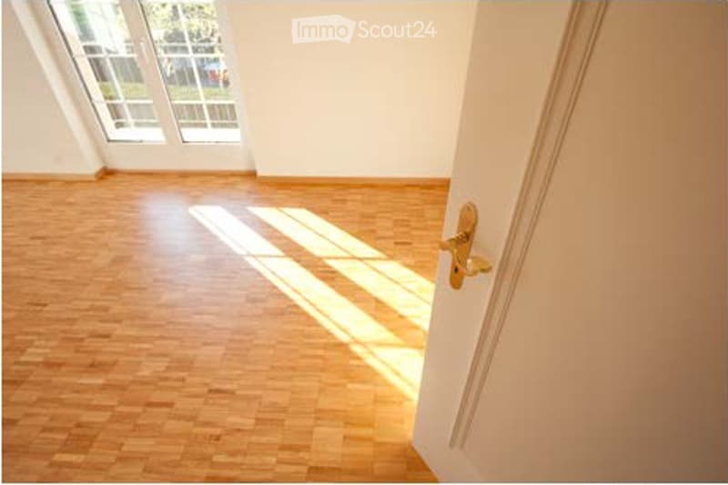 Empty room with wooden flooring, white walls, and large windows allowing natural light to enter. The room is unfurnished and appears to be a living space or bedroom.