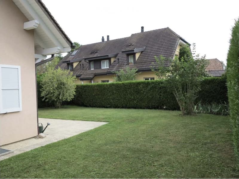 Well-maintained garden with a neatly trimmed lawn, a hedge border, and a small watering can near the house entrance.