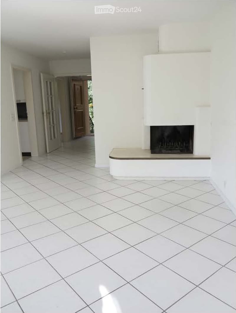 Empty living room with white tiled flooring, white walls, and a built-in fireplace with a white surround. The room has open doorways leading to other areas.