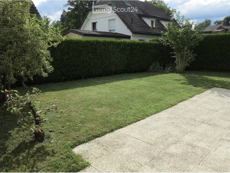 Well-maintained garden with a neatly trimmed lawn, a concrete pathway, and surrounding hedges and trees. A modern house is visible in the background.
