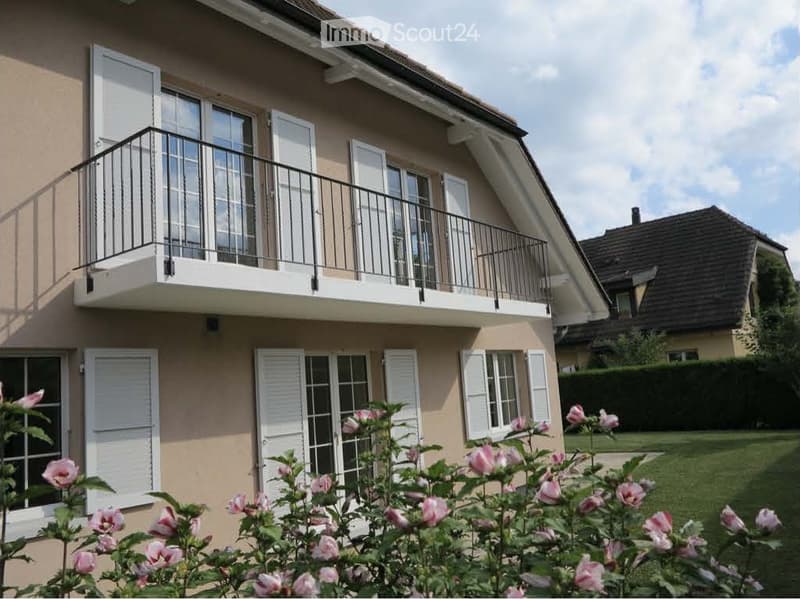 Two-story house with beige exterior, white shutters, and a balcony on the upper floor. Pink roses are in the foreground, and there is a well-maintained lawn.