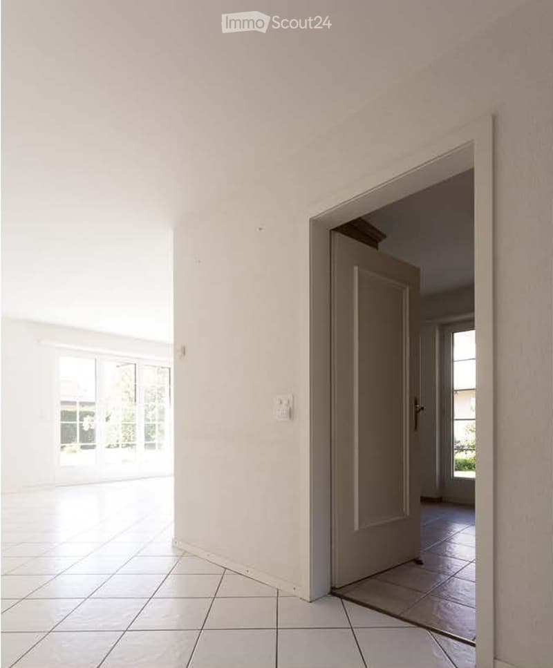 Empty room with white walls, tiled floor, and large windows. A door leads to another room with tiled flooring.