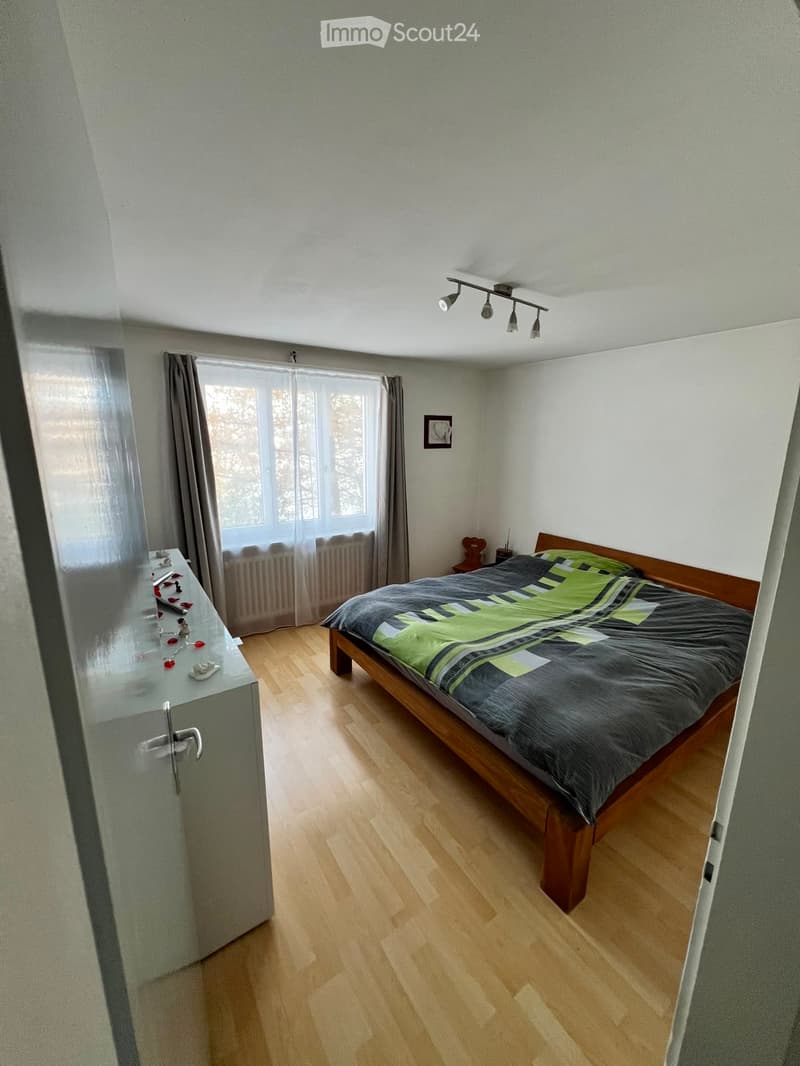 Bedroom with wooden floor, large window with curtains, bed with green and black bedding, nightstand, and cabinet.