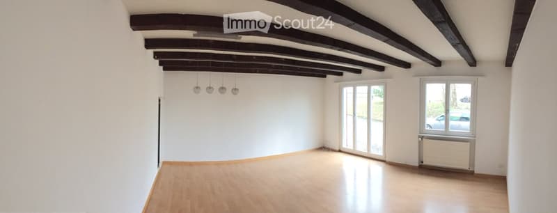 Empty room with wooden flooring, white walls, exposed wooden beams on the ceiling, and large windows with blinds. Natural light fills the space.