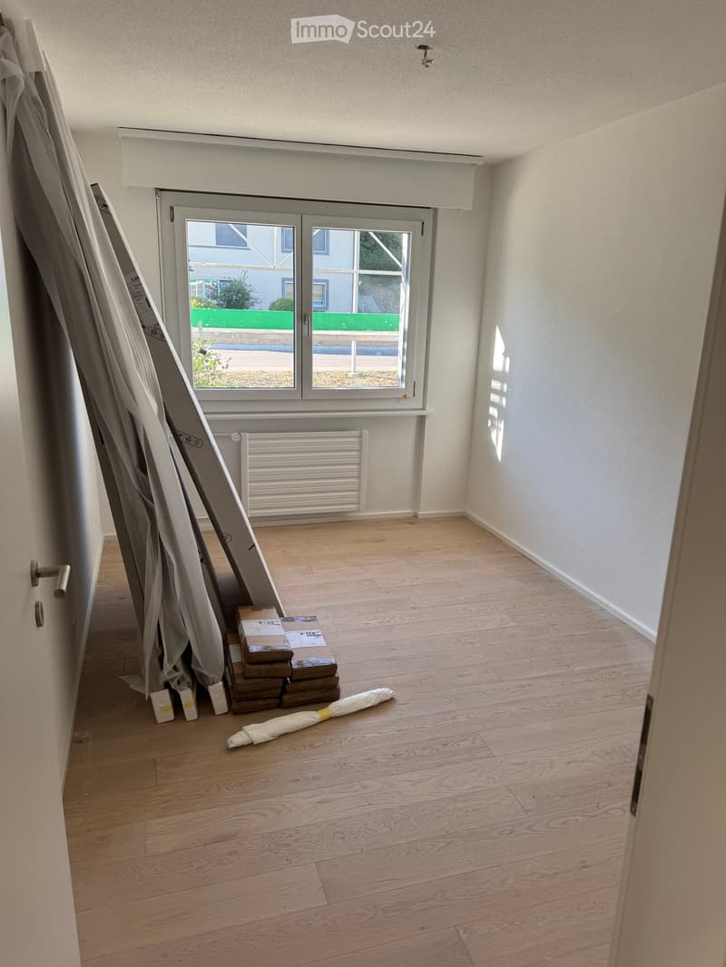 Empty room with wooden flooring, white walls, large window with blinds, radiator, and some stacked boxes and rolled-up items on the floor.