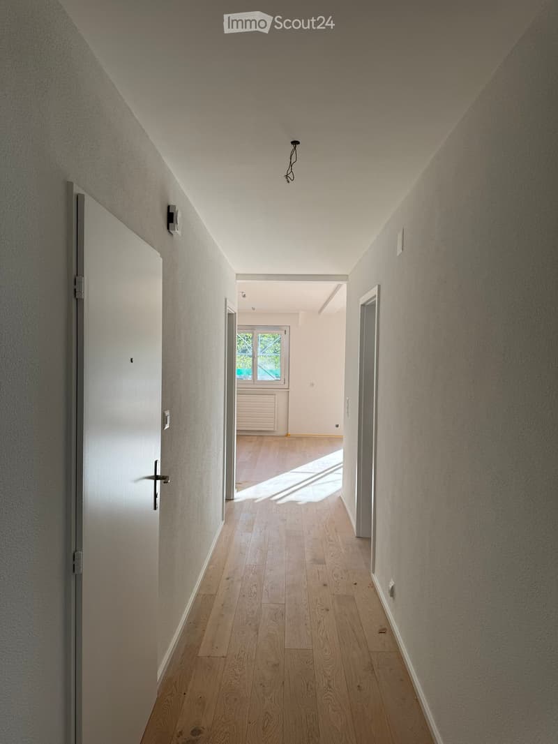A narrow hallway with wooden flooring, white walls, and a slanted ceiling. Natural light streams in from a window at the end, illuminating the space.