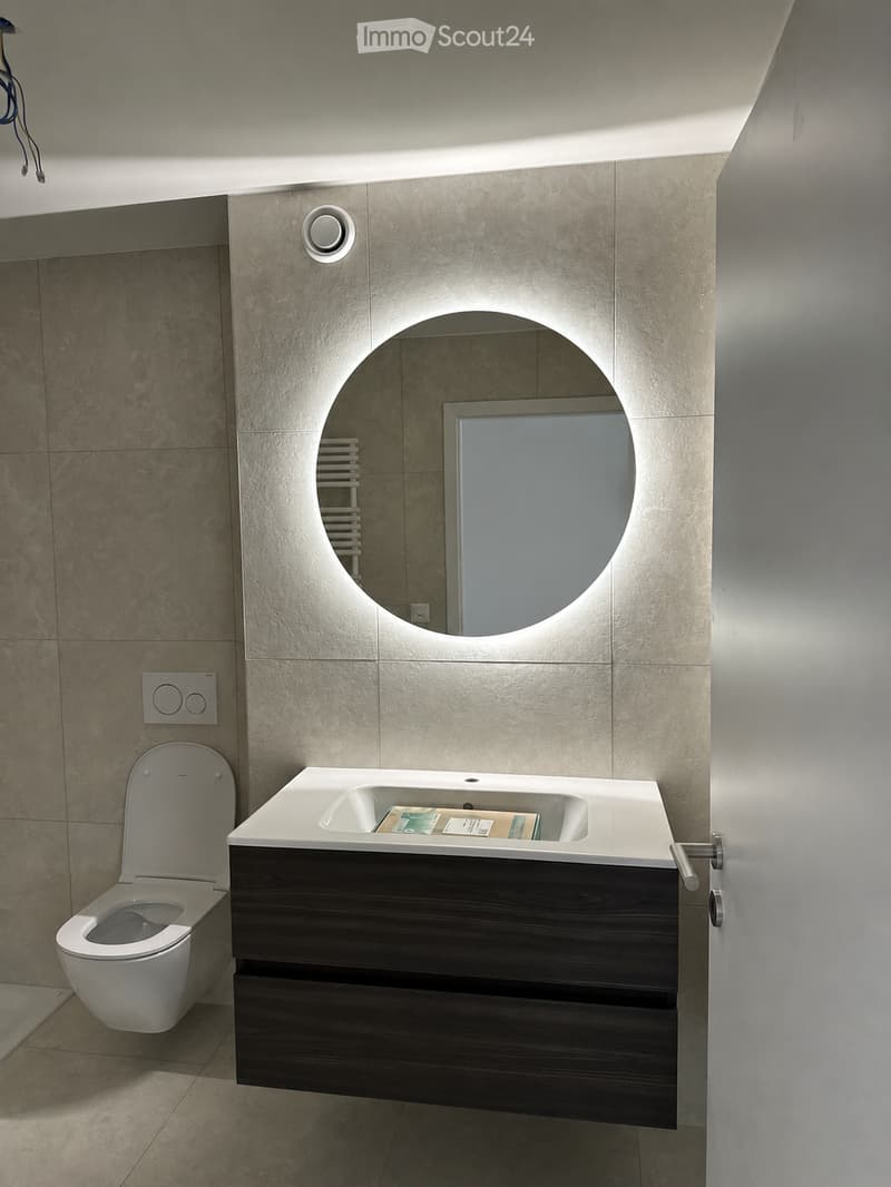 Modern bathroom with a white toilet, dark wooden sink cabinet, and a large circular mirror with integrated lighting.
