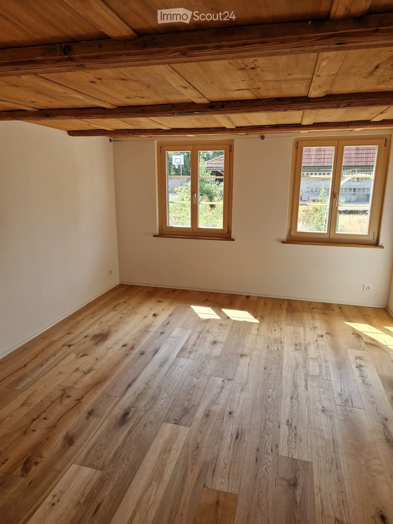Empty room with wooden floor and exposed wooden beams on the ceiling. Two windows with wooden frames provide natural light.