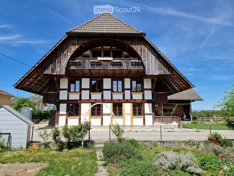 Traditional wooden house with white and brown facade, large arched balcony, multiple windows, and surrounding garden area.