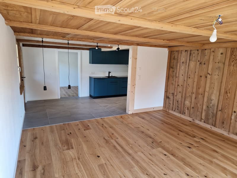 Empty room with wooden flooring, exposed wooden ceiling beams, and a small kitchen area with dark cabinets and a sink.