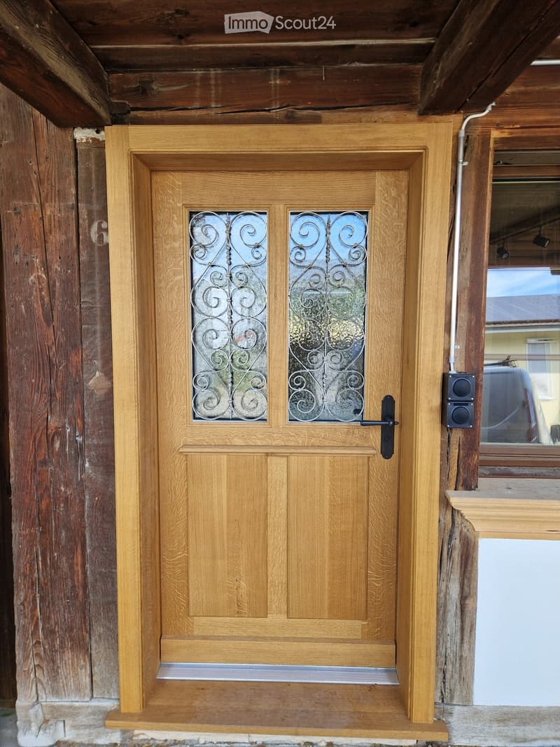 Wooden front door with decorative glass panels and a small step leading up to it, flanked by wooden beams and a window to the right.