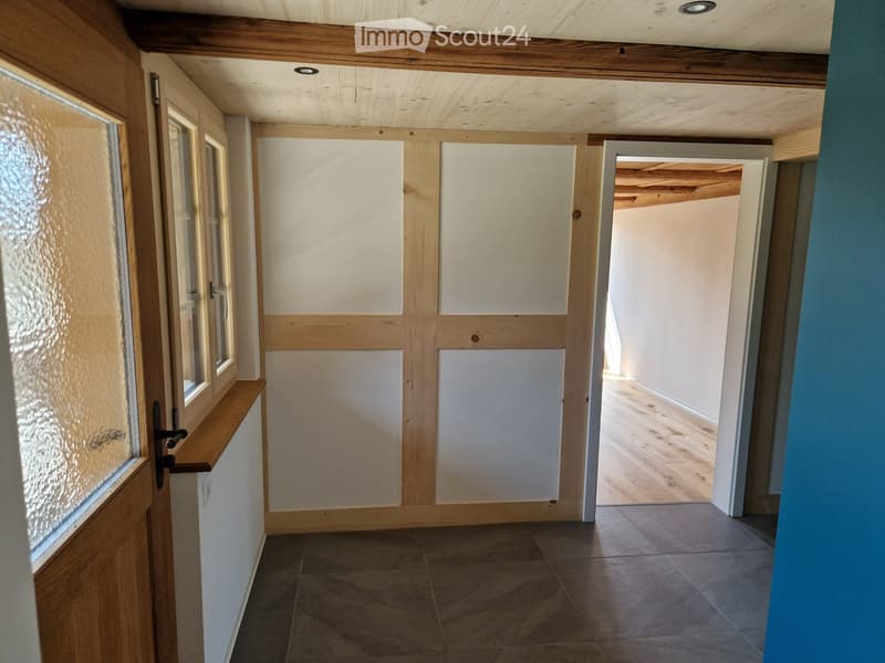 Empty hallway with wooden ceiling, tiled floor, and white walls. Features a frosted glass window on the left and an open doorway leading to another room with wooden flooring.