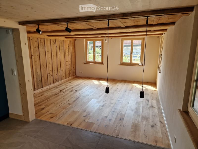 Empty room with wooden floor, exposed wooden ceiling beams, and two hanging pendant lights. Natural light from two windows on the far wall.
