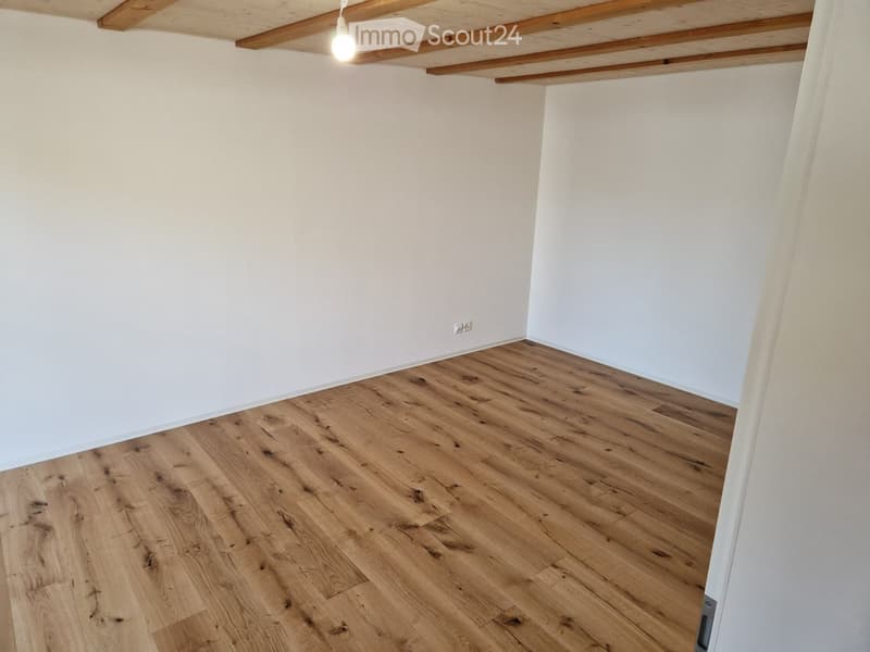 Empty room with light wooden flooring and white walls, exposed wooden ceiling beams, and a single light fixture.