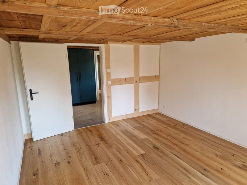 Empty room with wooden flooring, white walls, and a wooden ceiling. A white door is open to another room with dark blue cabinets.