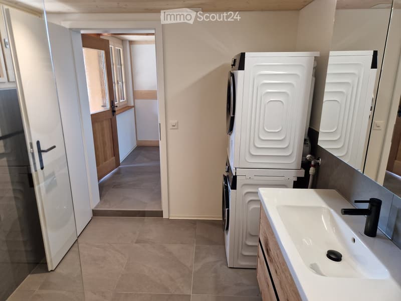 Modern bathroom with a white sink, black faucet, washing machine, and a large mirror. The floor is tiled, and there is an open doorway leading to another room.