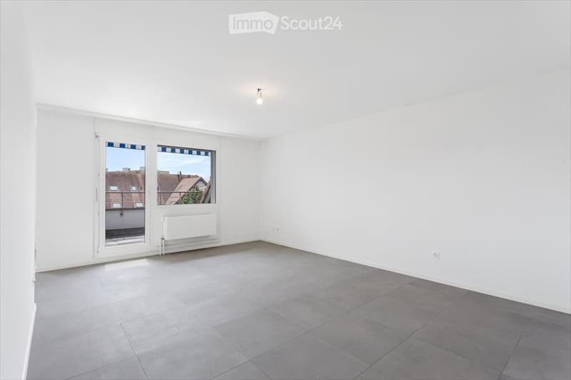 Empty room with white walls, grey tiled floor, large window with blinds, radiator, and single ceiling light fixture.