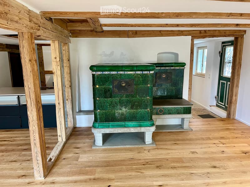 Empty room with wooden floor, exposed wooden beams, and a green stove with decorative tiles.