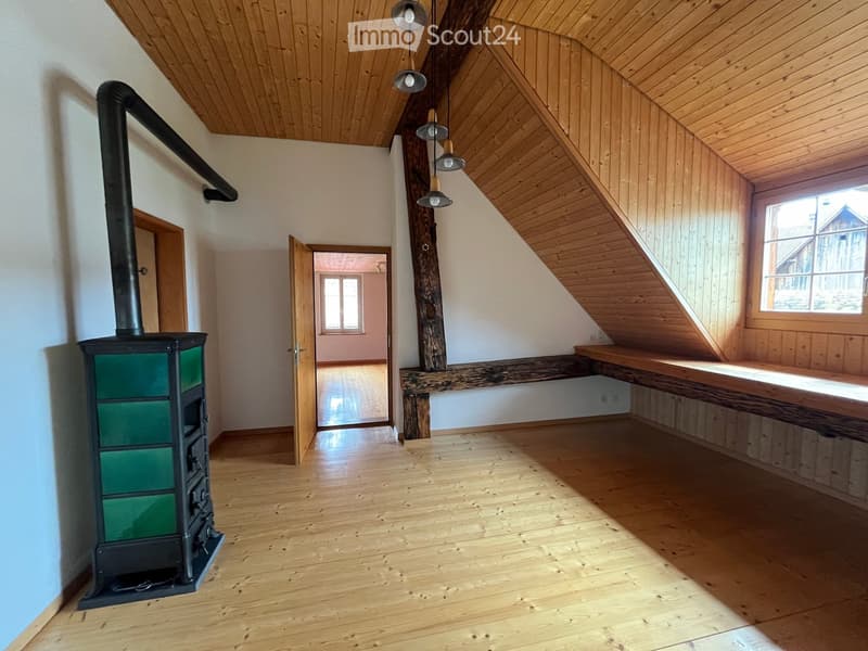 An empty attic space with wooden floors, white walls, and exposed wooden beams. Features a black wood-burning stove with a chimney, a built-in wooden bench, and large windows allowing natural light.