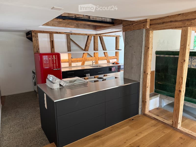 Modern kitchen with stainless steel countertop, black cabinets, red refrigerator, and exposed wooden beams. Includes a sink, faucet, and washing machine visible.