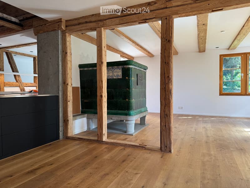 An unfurnished room with exposed wooden beams and polished wooden flooring, featuring a large green fireplace and a window with wooden frames.