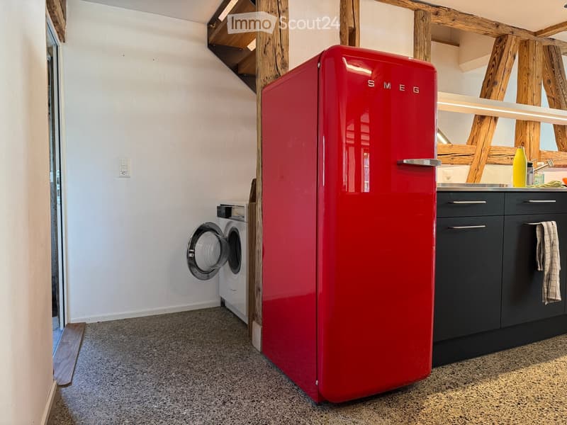 A modern kitchen with a red vintage-style refrigerator, dark grey cabinets, a washing machine, and a small sink area with a bottle on the counter.