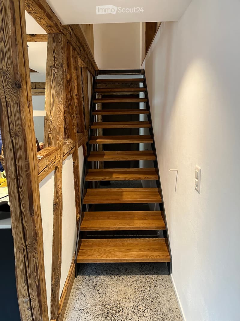 Wooden staircase with a modern design, featuring a curved wooden handrail and light-colored wooden steps, set against white walls and a textured concrete floor.
