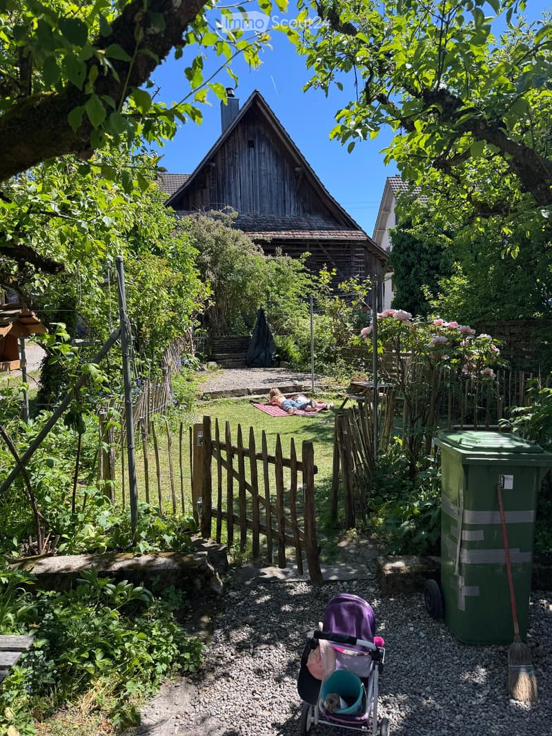 Garden area with a wooden fence, a small wooden shed, a grassy lawn, and a picnic table. A stroller is visible in the foreground, and a person is lying on a blanket in the background.