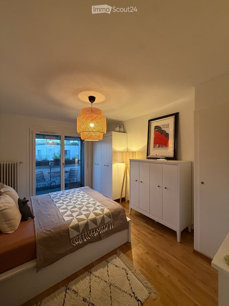 Modern bedroom with a bed, geometric quilt, white cabinets, pendant light, and a framed picture on the wall. The room has wooden flooring and a white shag rug.