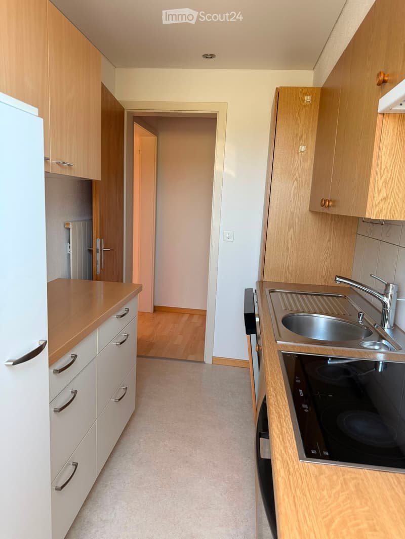 Modern kitchen with light wooden cabinets, stainless steel sink, and built-in stove. The space features a clean, minimalist design with neutral tones and a visible doorway leading to another room.