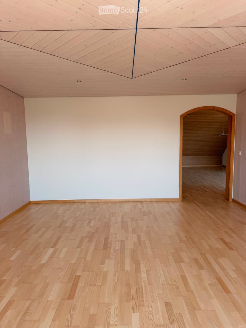 Empty room with light wooden flooring and white walls, featuring a wooden ceiling with geometric patterns and an arched doorway leading to another space.