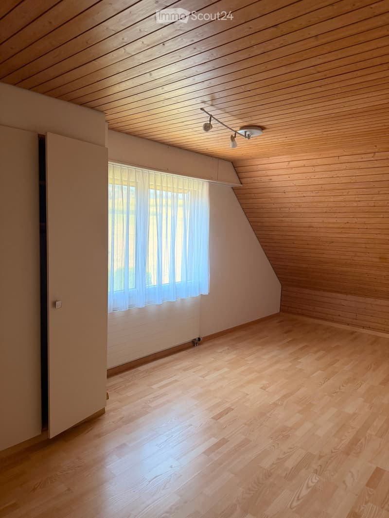 Empty attic room with wooden walls, ceiling, and floor, featuring a window with sheer curtains and a track light fixture.